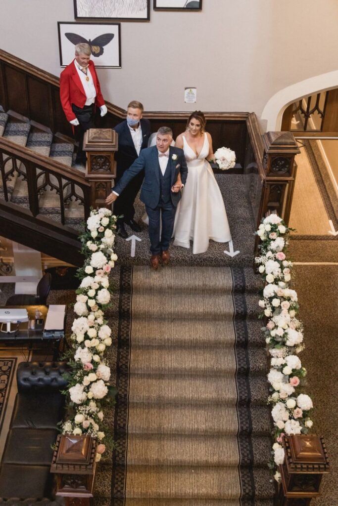 46 bridal party decends flower decked staircase de vere hotel wotton under edge oxfordshire wedding photoghers